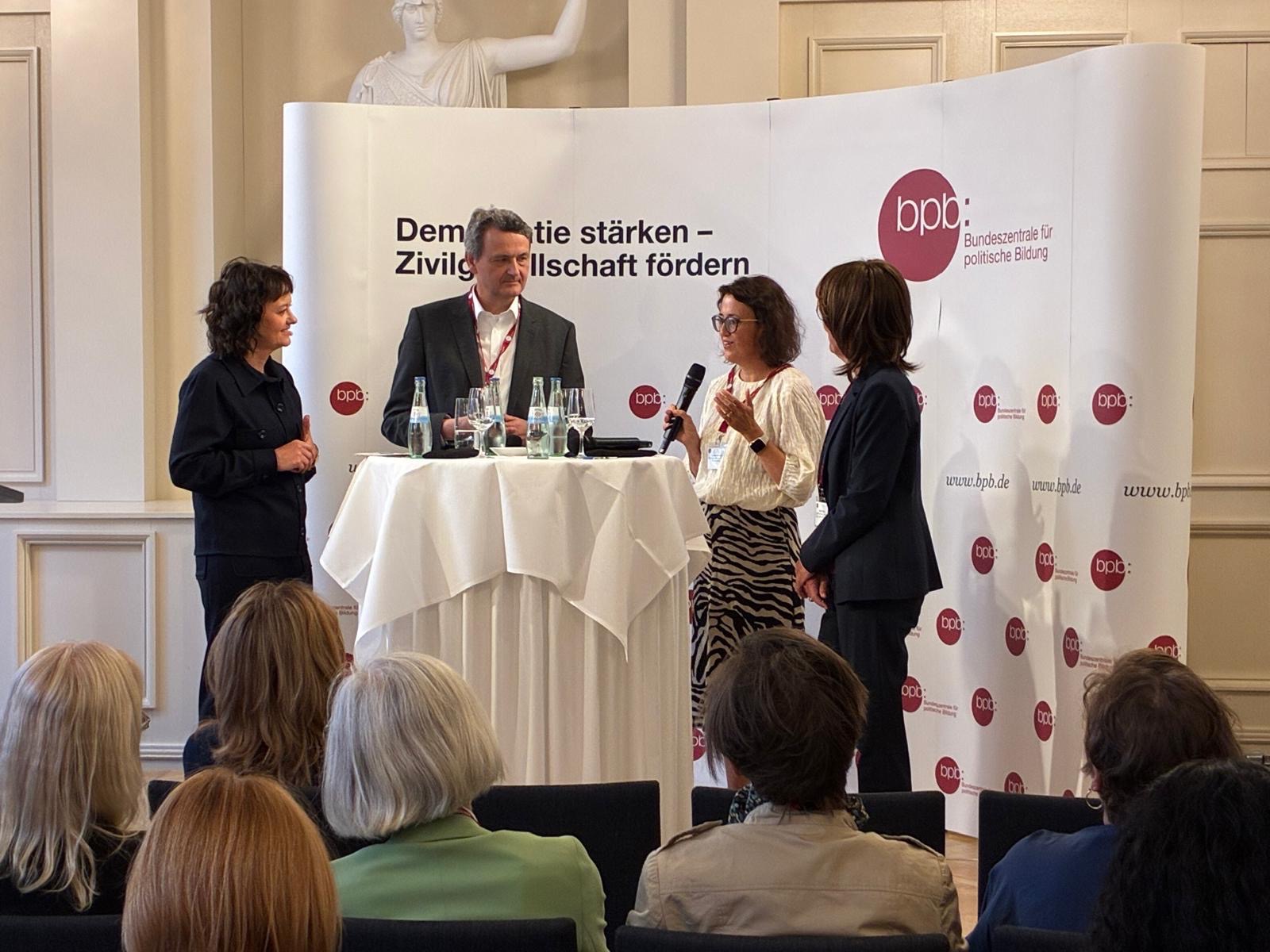 Andrea Rühmann und weitere Speaker*innen stehen auf einem Podium
